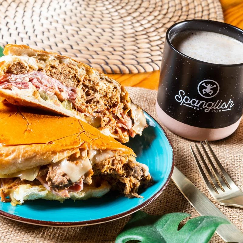 Kokí Cubano sandwich cut in half on a turquoise plate beside a cup of coffee