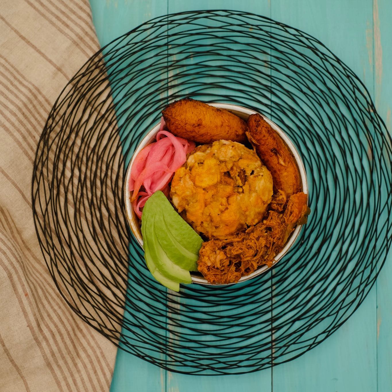 Kokí El Coquí mofongo bowl with shredded pork, sweet plantains, avocado, and pink onions on a turquoise table